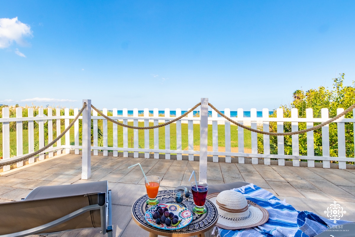 The outdoor terrace features a low table adorned with fresh fruits and colorful beverages. A lounge chair is positioned nearby, with a wide view of the sea beyond a white picket fence. A blue and white striped blanket rests on the ground, enhancing the relaxing ambiance.