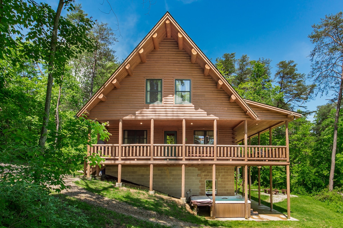 The exterior of the lodge is showcased, highlighting its wooden structure and slanted roof. A spacious porch runs along the front, complete with railings, providing access to the hot tub below. Surrounding greenery and trees complement the natural setting of the lodge.