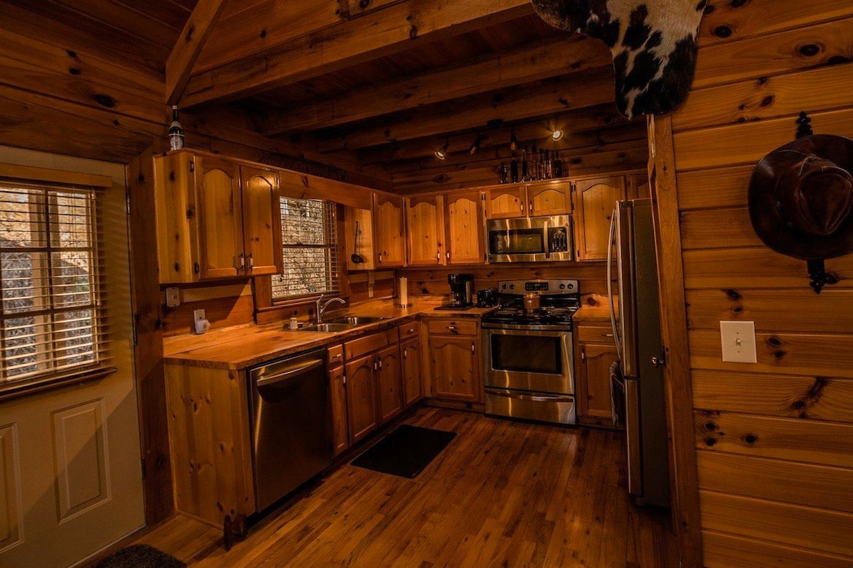 The kitchen features natural wood cabinetry and countertops, with stainless steel appliances including a stove, refrigerator, and dishwasher. Ample natural light enters through windows adorned with wooden blinds. A warm wooden floor complements the rustic design of the space.