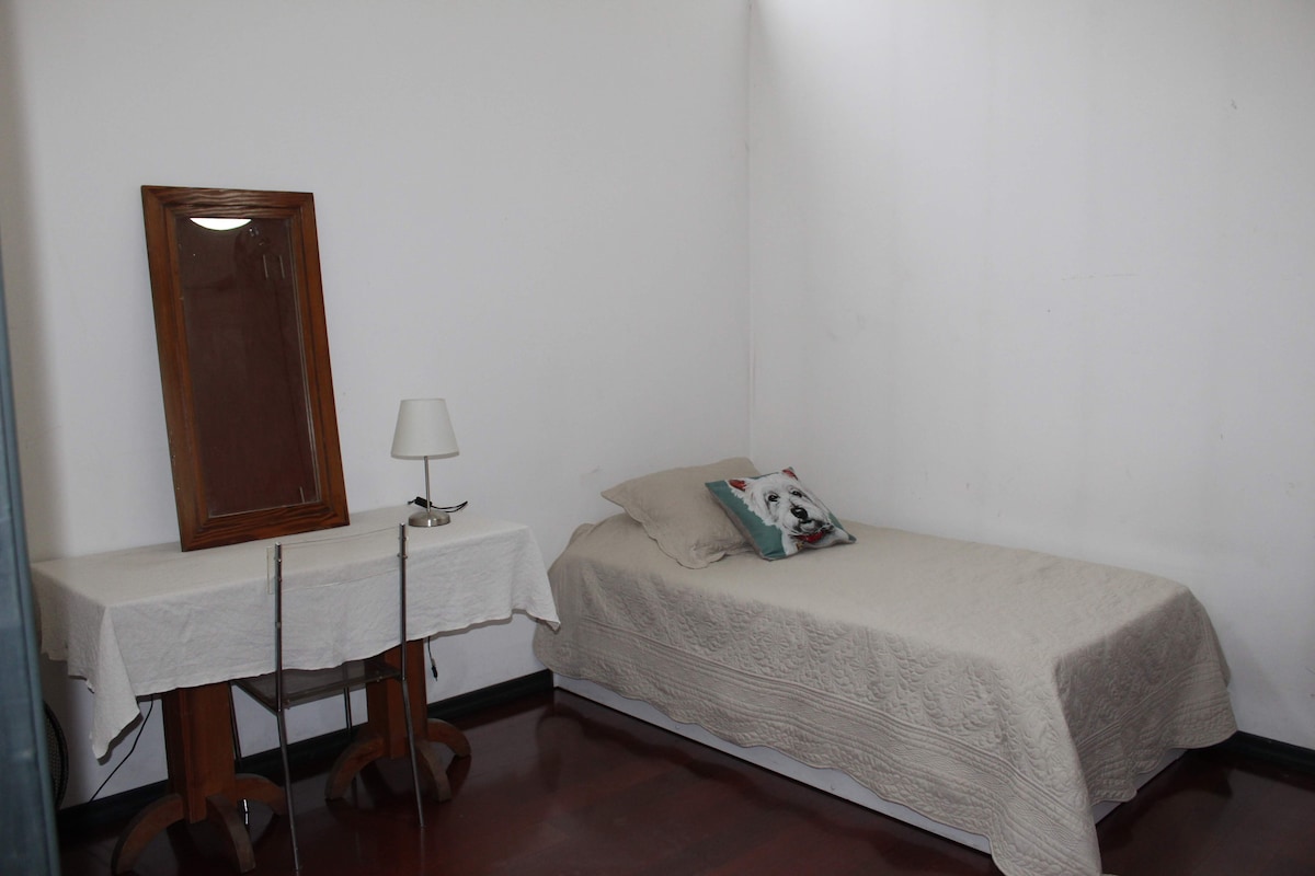 A simple bedroom features a single bed covered with a light-colored blanket. A small table with a table lamp stands beside the bed, and a mirror is mounted on the wall. Natural light enters from a window, enhancing the room's spacious feel.