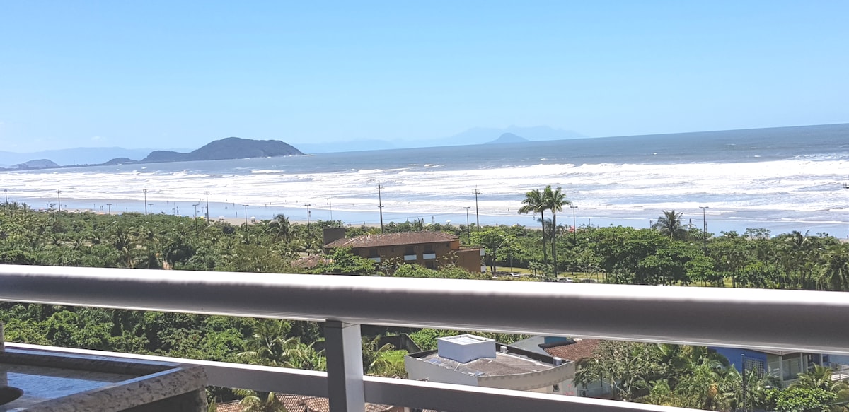 A clear view of the expansive sea is captured from the balcony. Rolling waves can be seen meeting the shoreline, framed by lush greenery and distant hills on the horizon. Natural light enhances the serene atmosphere, inviting relaxation.