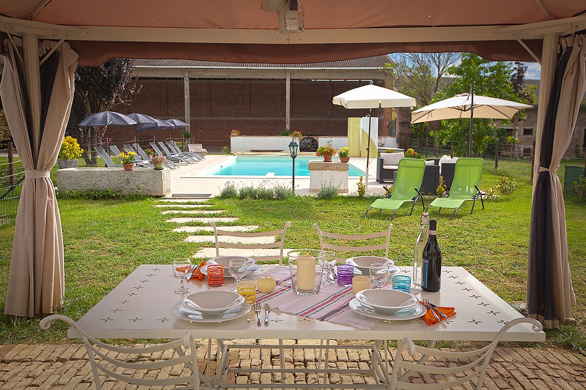 A shaded outdoor dining area features a white table set with plates and glasses, overlooking a well-maintained garden. In the background, a sparkling pool is surrounded by lounge chairs and umbrellas, creating an inviting atmosphere for relaxation and enjoyment.