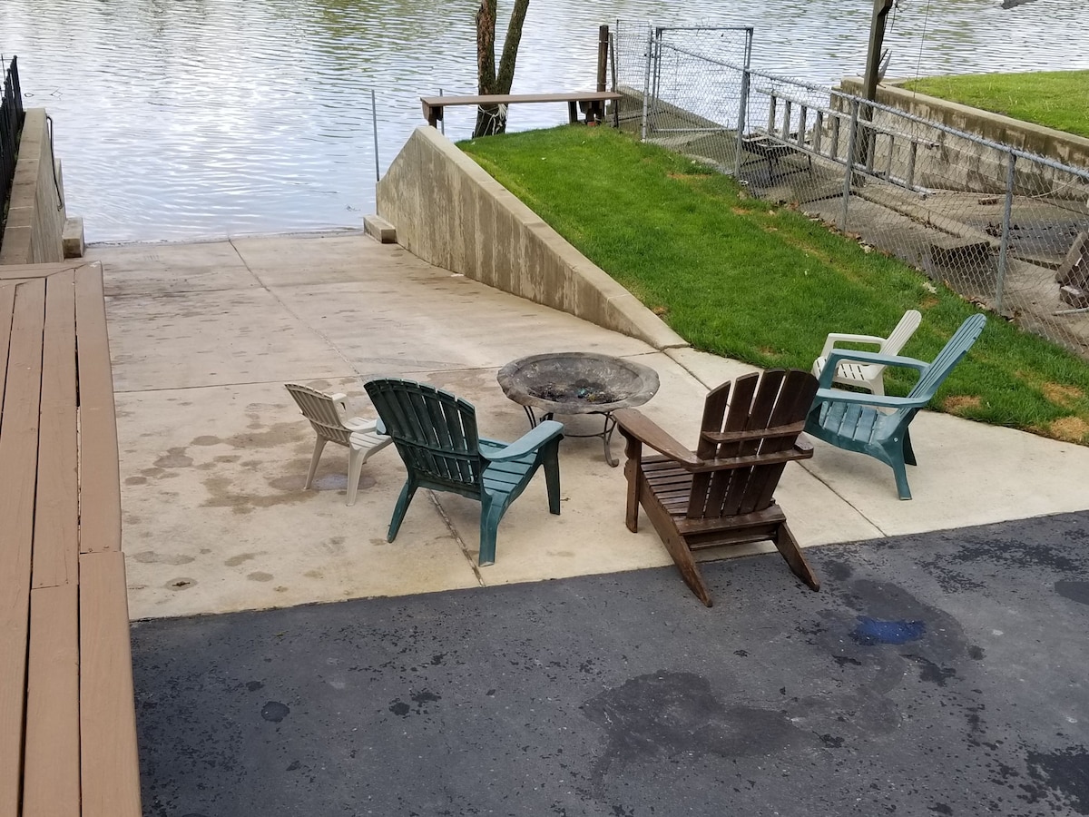 A fire pit area is designated by several adirondack chairs arranged around it, creating an inviting space for gatherings. A grassy slope leads down to the waterfront, with a concrete ramp visible beside it, providing access to the water.