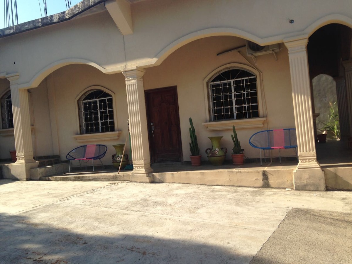 The exterior features a structured facade with arched doorways and decorative columns. Two chairs in light blue and pink sit on a concrete porch, complemented by potted plants. The entrance is marked by a wooden door, framed by large windows.