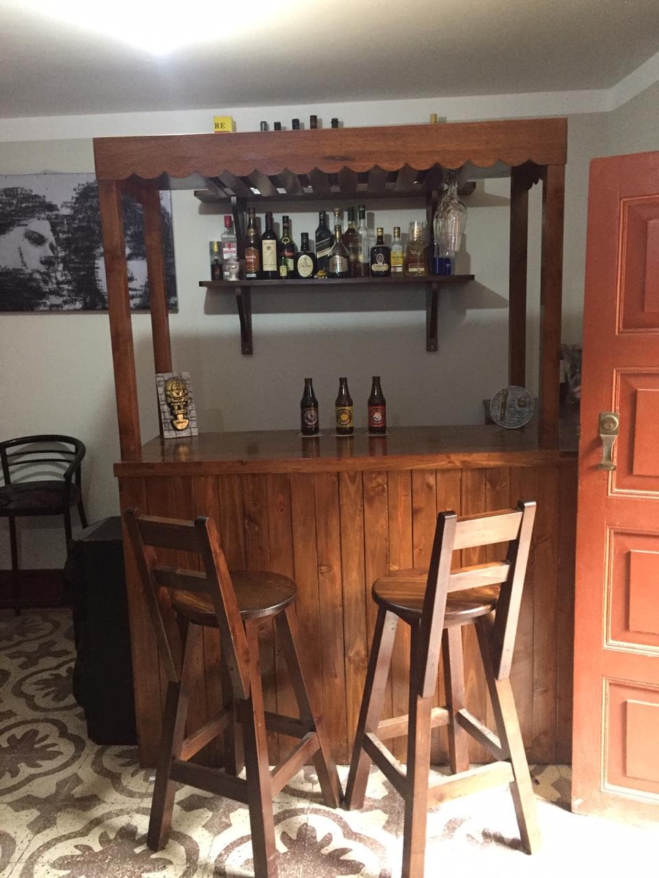 A wooden bar with a polished countertop is showcased, featuring shelves stocked with various bottles. Two high stools are positioned in front, inviting guests to sit. The patterned floor adds texture to the area, complementing the warm wood tones.