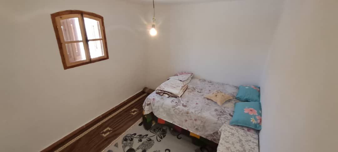 A simple bedroom features a colorful bed with a floral-patterned duvet and several light pillows. A small bedside area provides additional space for personal items. Natural light enters through a wooden window with shutters, illuminating the room's white walls and wooden flooring.