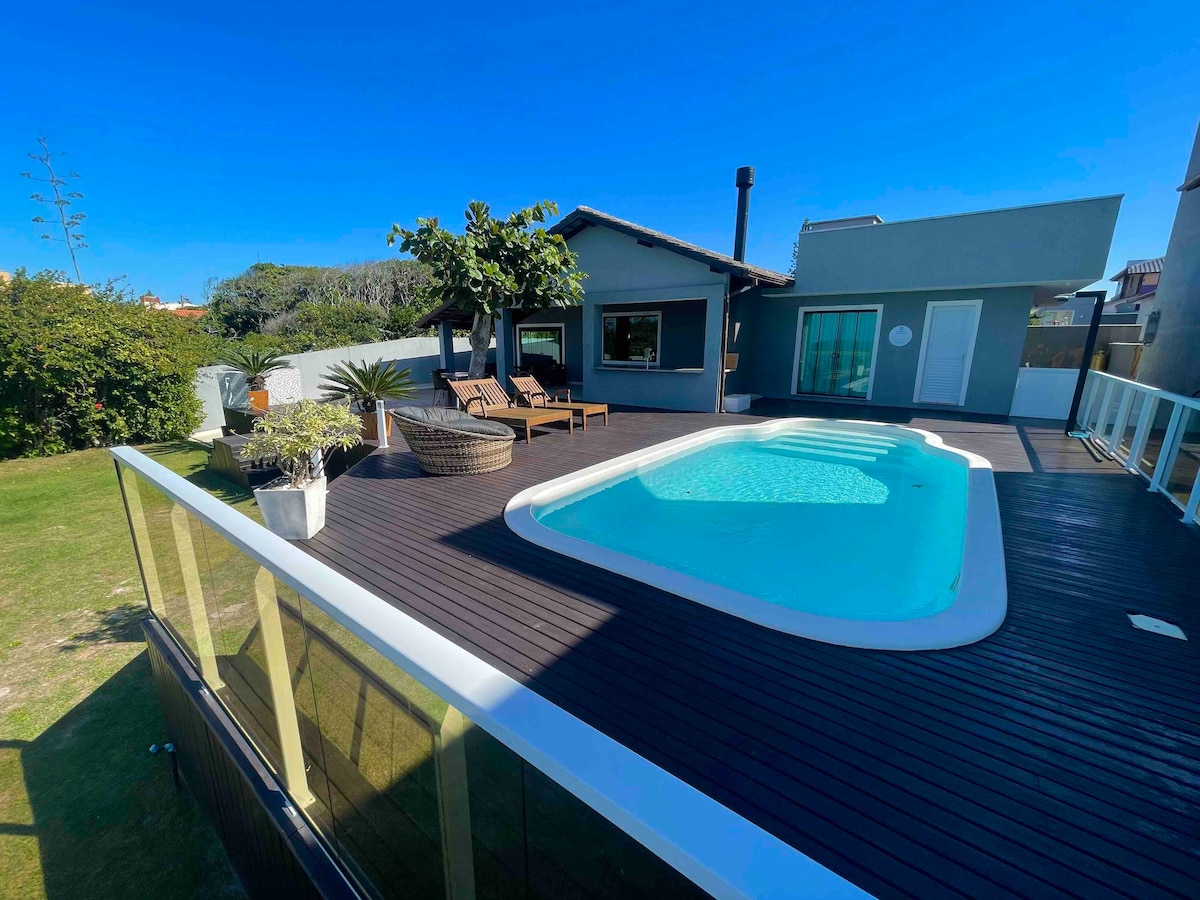 An outdoor area is featured with a private pool surrounded by a wooden deck. Lounge chairs are positioned nearby, offering a space for relaxation. Lush green landscaping and a single tree add a natural element to the setting, under a clear blue sky.
