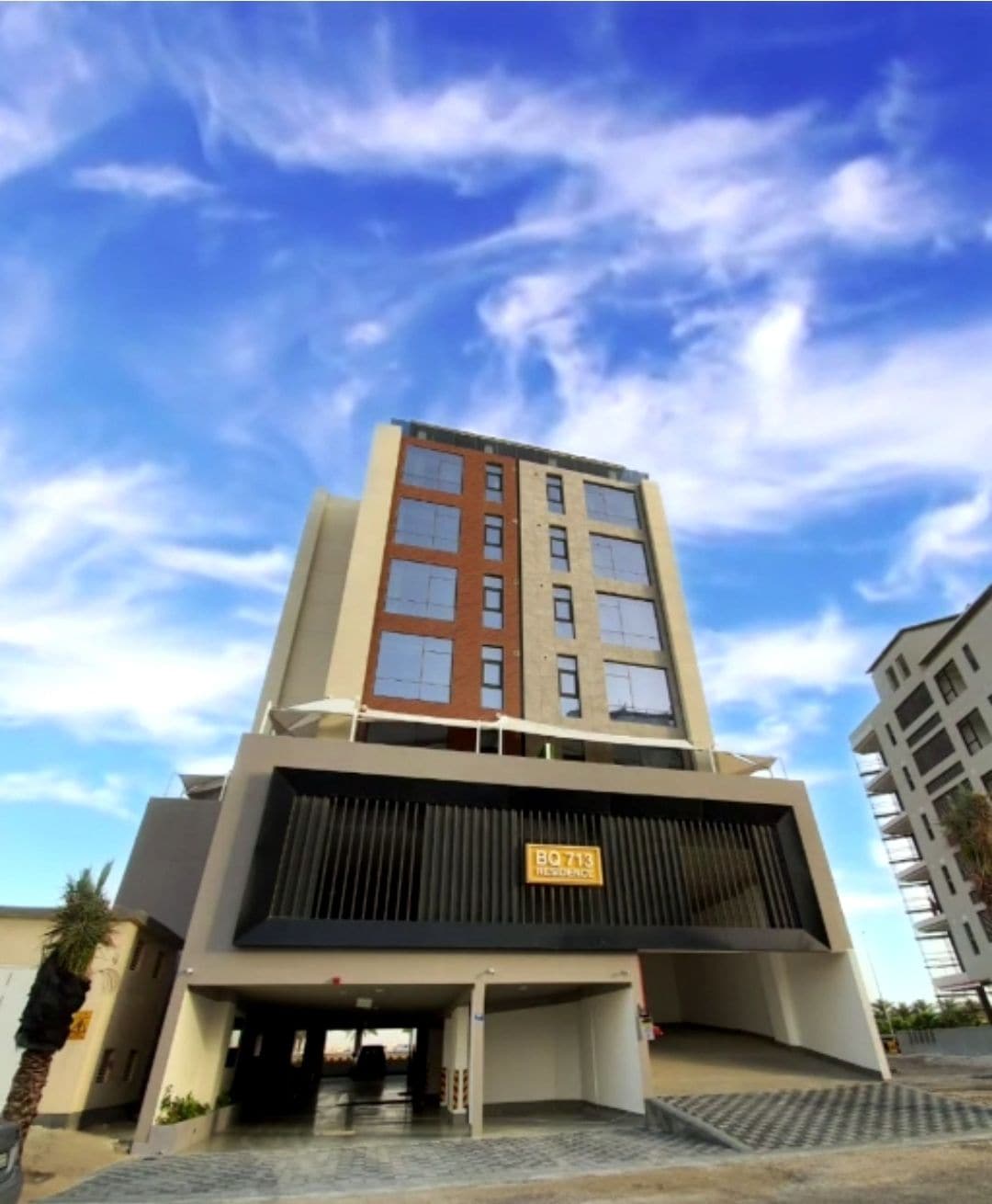 The exterior view of the building showcases a modern design with a combination of dark and light facade elements. Clear blue skies and white clouds provide a bright backdrop, while palm trees are visible at the sides, enhancing the urban landscape.