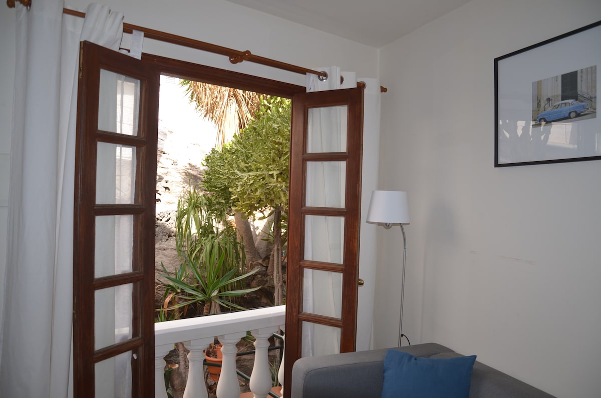 A set of wooden French doors opens to a small balcony, adorned with lush greenery. The interior features a comfortable sofa with a blue accent pillow. A tall floor lamp is positioned beside the sofa, adding to the simplicity of the room.