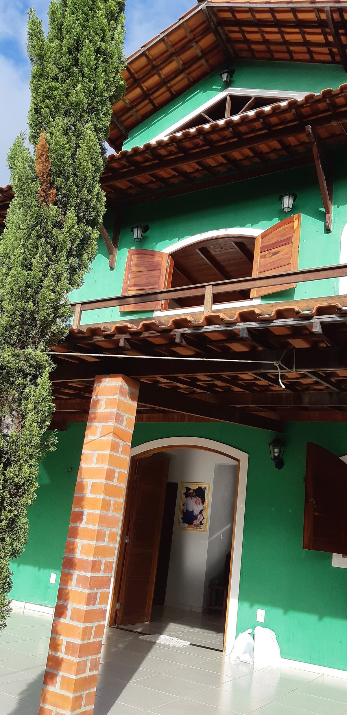 The exterior of a green house is visible, featuring a brick pillar supporting a covered porch. Wooden shutters adorn the upper floor windows, allowing natural light to filter through. Tall cypress trees flank the entrance, adding to the inviting atmosphere.