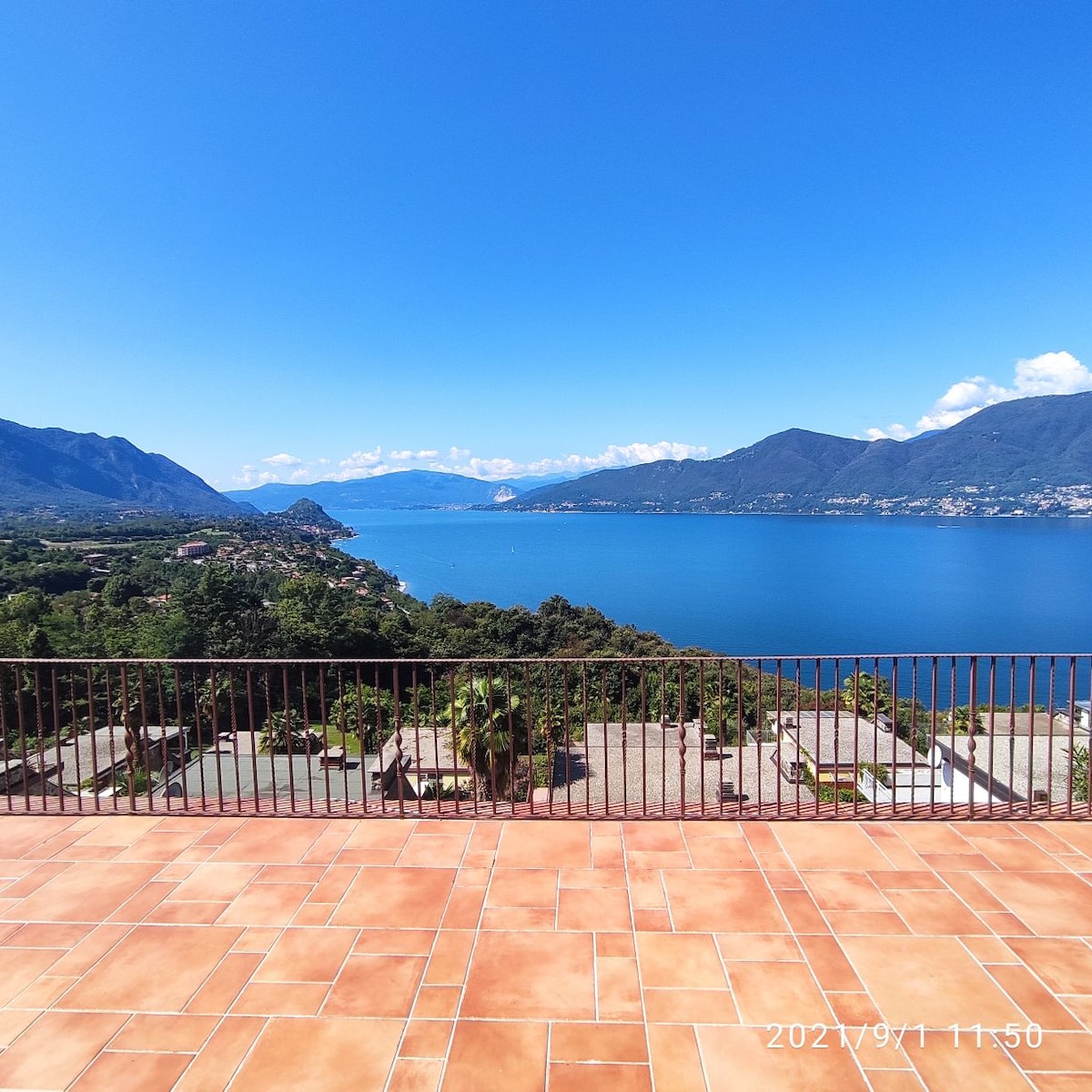 An expansive terrace showcases a sweeping view of the lake and surrounding mountains, accentuated by a clear blue sky. The sunlit area features terracotta tiles, creating a spacious and inviting outdoor setting.