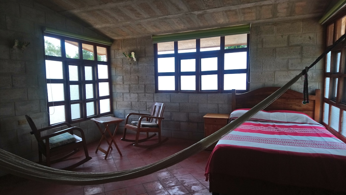 A well-lit room is furnished with a spacious bed and a hammock. Two large windows provide natural light, while a small table and two chairs are positioned by the window. The walls are made of stone, adding a rustic touch.