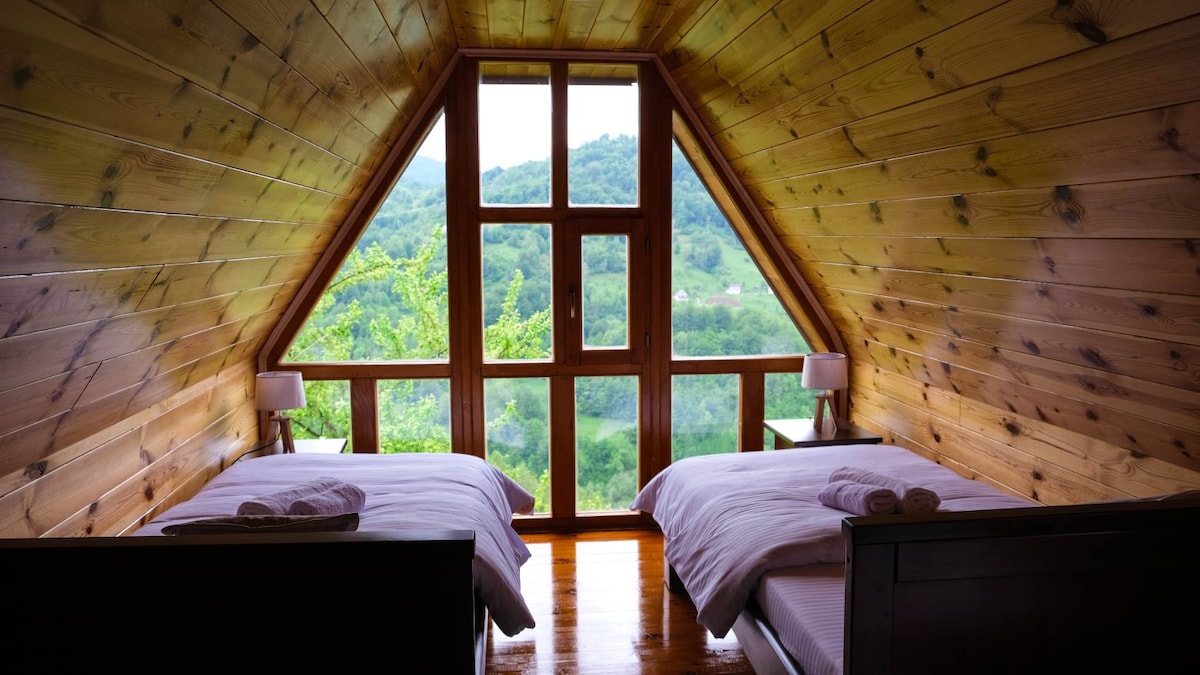 An airy room features two spacious beds with neatly arranged towels. Large windows frame scenic views of the surrounding greenery, while warm wooden paneling creates a cozy ambiance.