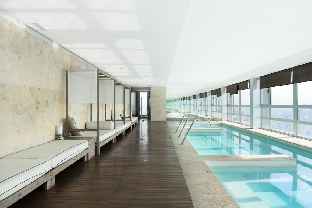An indoor pool area features large windows allowing natural light to fill the space. Sun loungers are positioned along the wooden floor, with a clean swimming pool visible. The walls are neutral with light stone, providing a modern and serene environment.