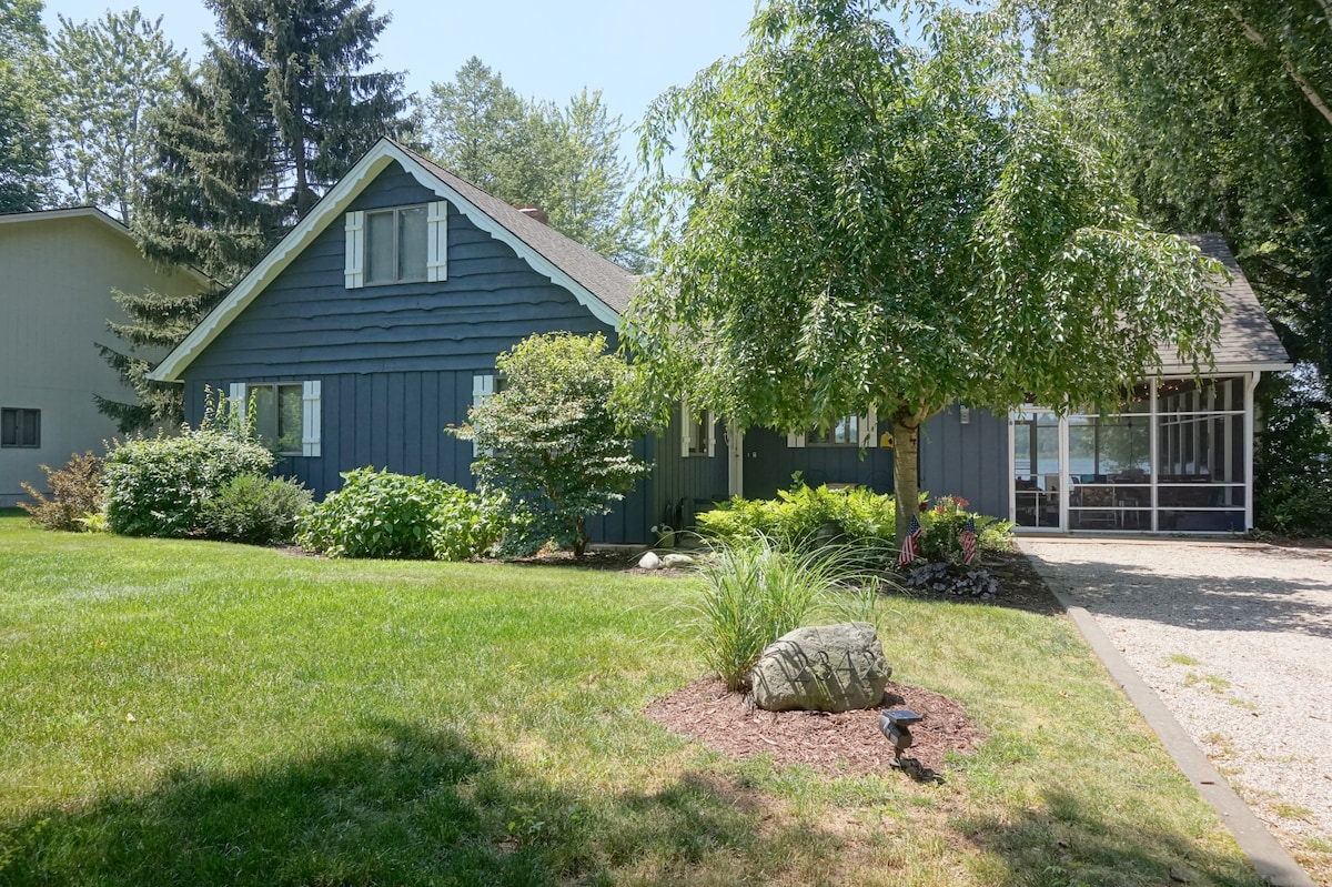 A charming cottage exterior is framed by a lush green lawn, dotted with bushes and trees. A gravel path leads to the entrance, with a screened-in porch visible on the right side. Natural light brightens the inviting facade of the home.