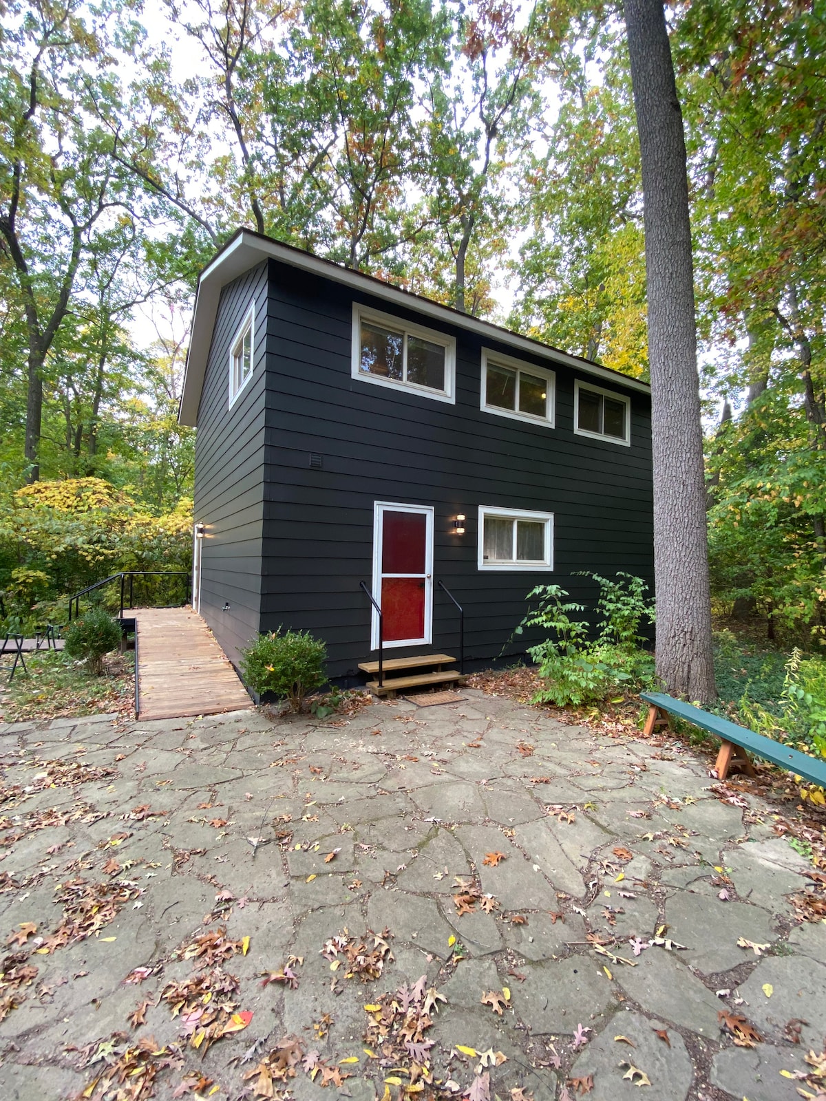 The exterior of the mid-century loft features a dark wood panel facade, complemented by large windows that invite natural light. A welcoming red door stands at the entrance, with stone pathway leading to a wooden deck surrounded by greenery and autumn leaves.