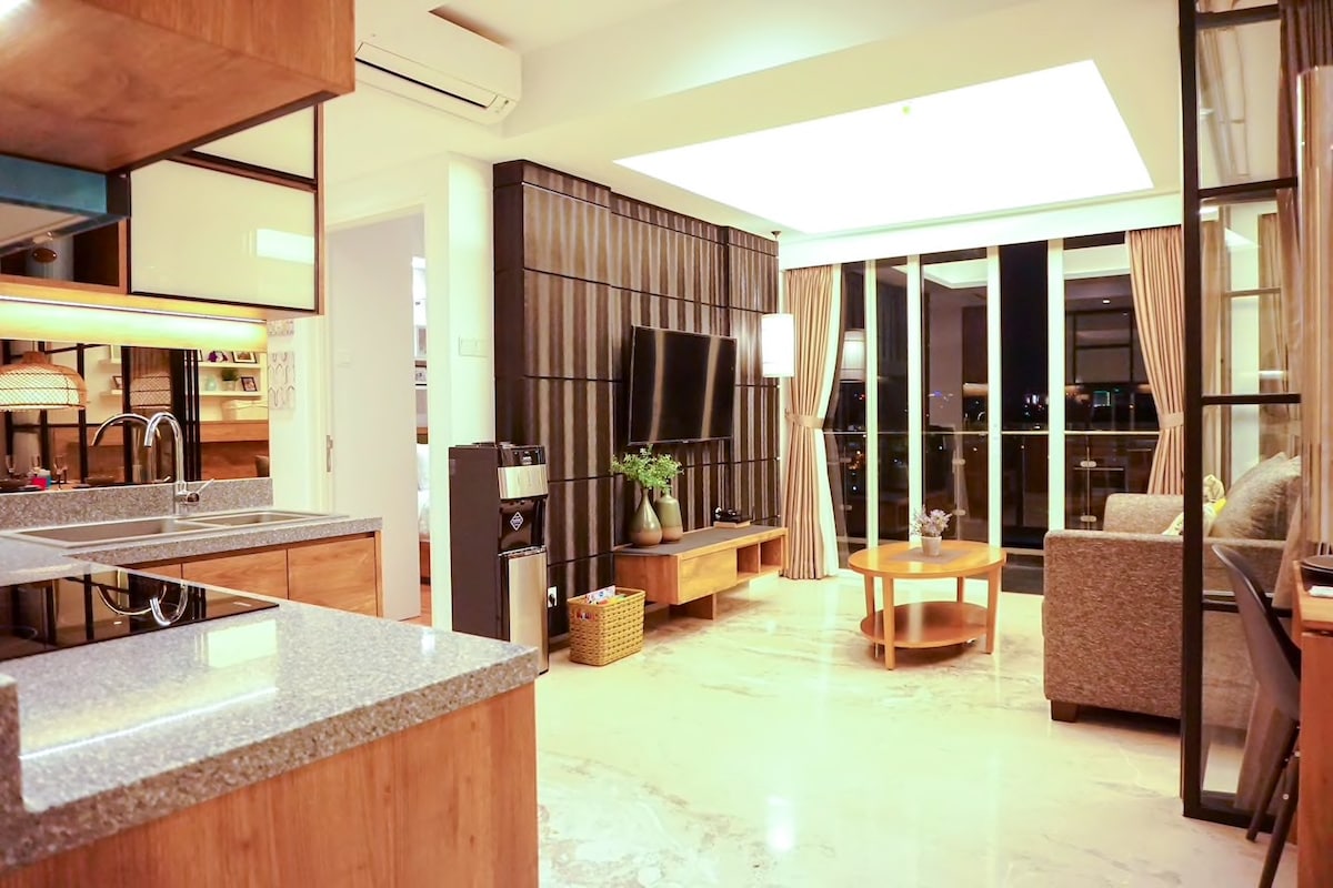 An open-concept living area is visible, featuring a modern kitchen with granite countertops and sleek cabinetry. A comfortable seating area includes a sofa facing a wall-mounted television. Large glass doors lead to a balcony, allowing natural light to brighten the space and showcasing views of the city.