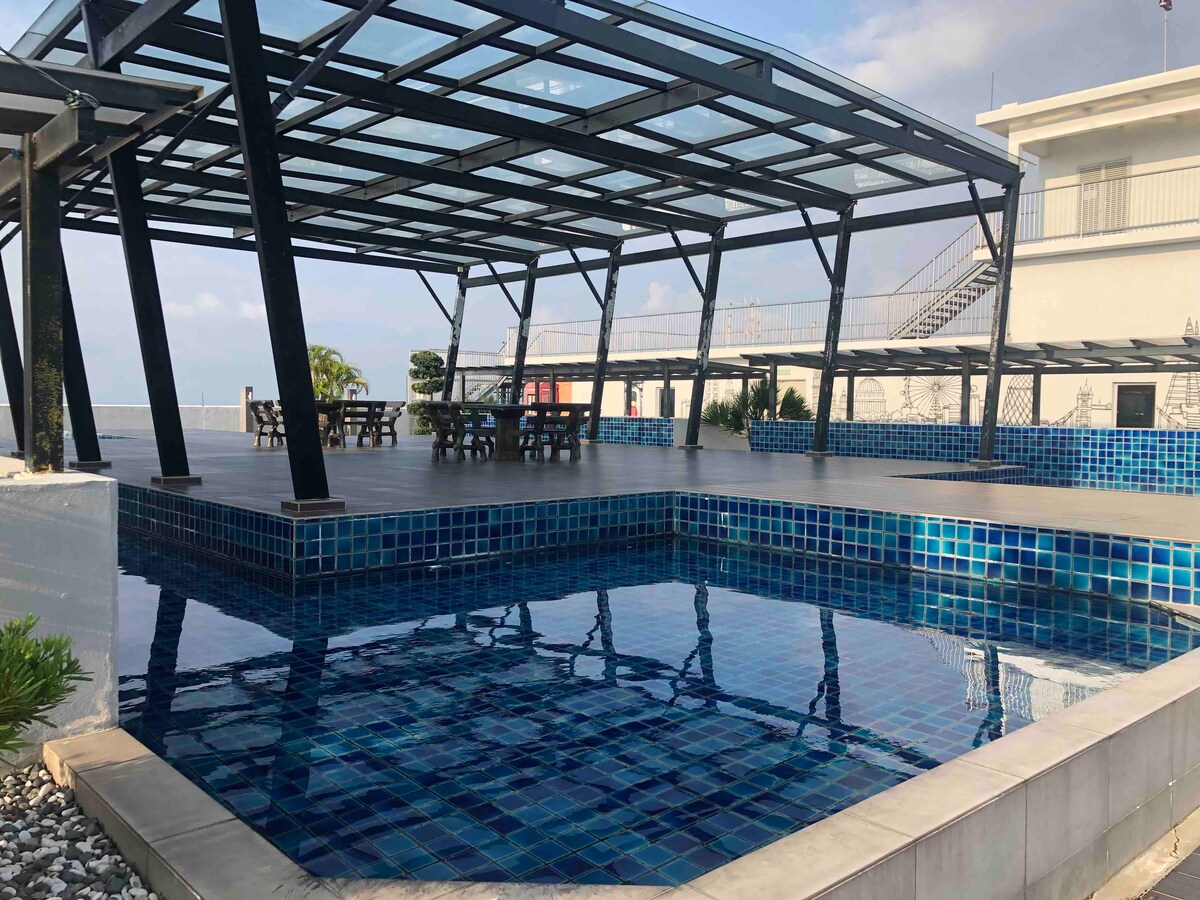 An outdoor area features a covered structure supported by metal beams, overlooking a swimming pool filled with blue tiles. Surrounding tables offer seating options. A tropical landscape is visible in the background, creating a serene environment for relaxation.
