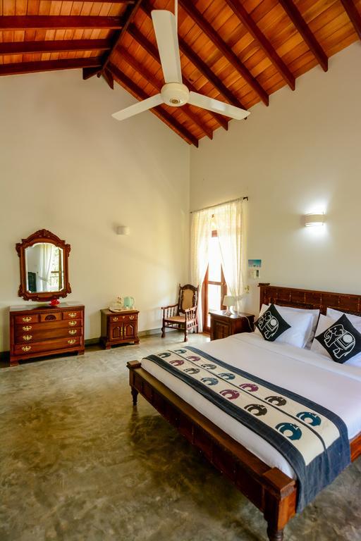 A spacious bedroom features high wooden ceilings and natural light filtering through the curtains. A large bed is centered against a decorative wall, complemented by two patterned bed cushions. A wooden dresser and a chair are positioned nearby, contributing to the room’s relaxed atmosphere.