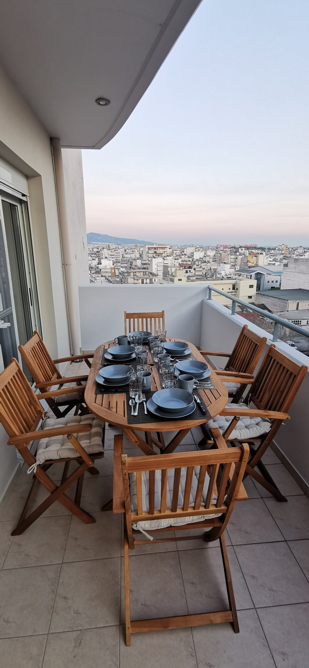 A wooden dining table is set for six, featuring plates and cutlery, on a balcony with a view of the city. Surrounding the table are comfortable folding chairs. The urban landscape extends in the background under a soft evening sky.