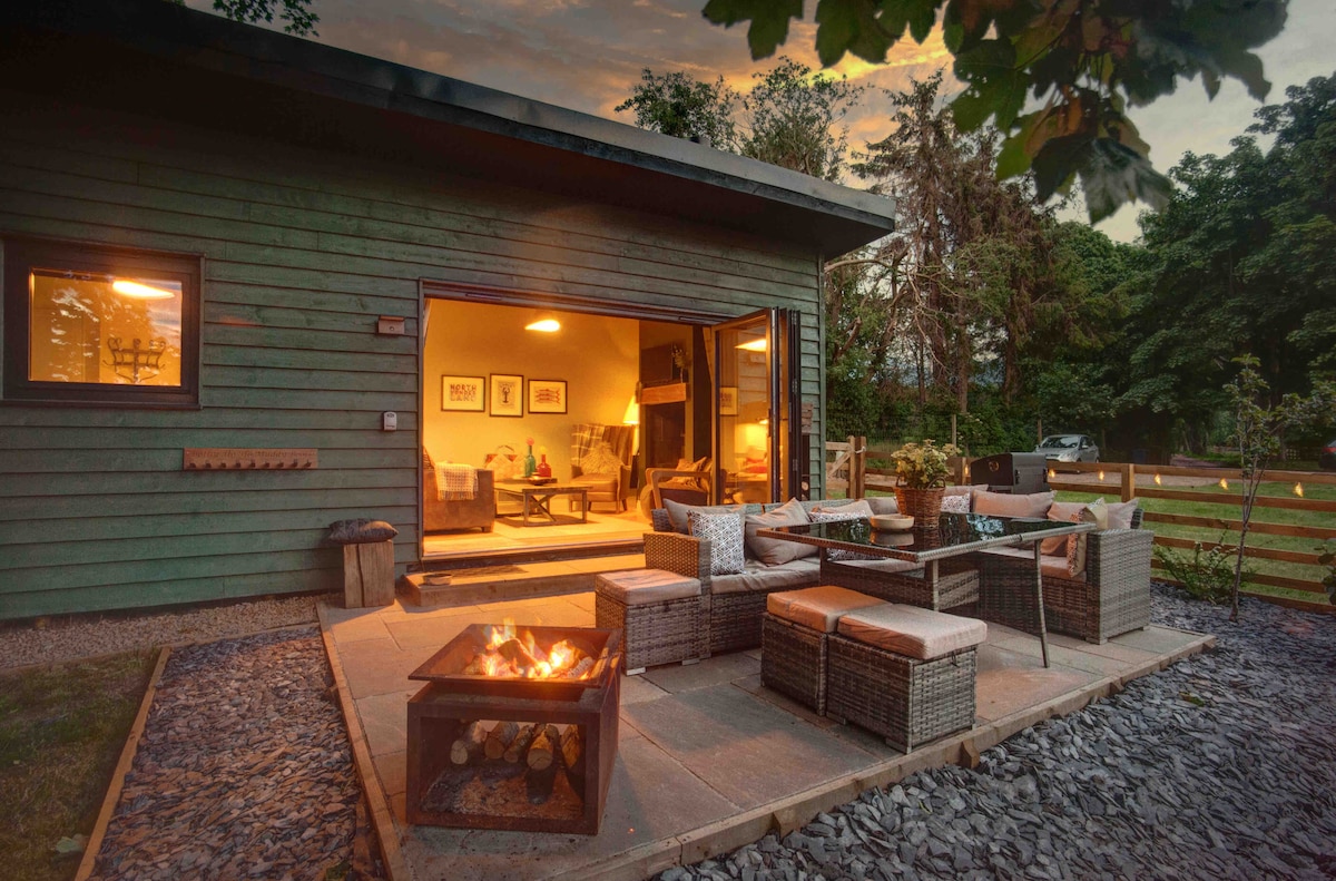 An outdoor seating area is displayed featuring comfortable wicker furniture surrounding a fire pit. Soft lighting from the building enhances the welcoming ambiance, while the natural stone patio complements the setting. The lush greenery of the garden provides a serene backdrop.