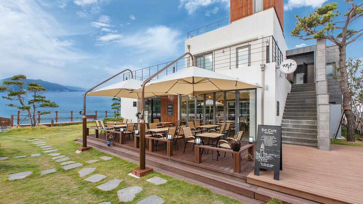 A spacious outdoor café area features multiple dining tables under large umbrellas. A wooden deck is complemented by a neatly manicured lawn and stone walkway. The serene ocean view and distant hills create a calming backdrop, inviting guests to relax in the open air.