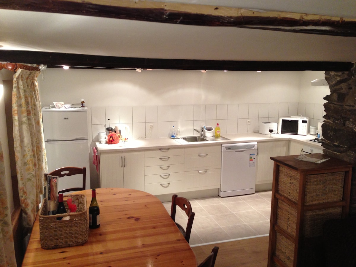 The kitchen area is equipped with modern appliances, including a fridge, dishwasher, and microwave. A wooden dining table with chairs is positioned nearby, accompanied by a basket of items. Soft lighting and white cabinetry enhance the clean and functional design.