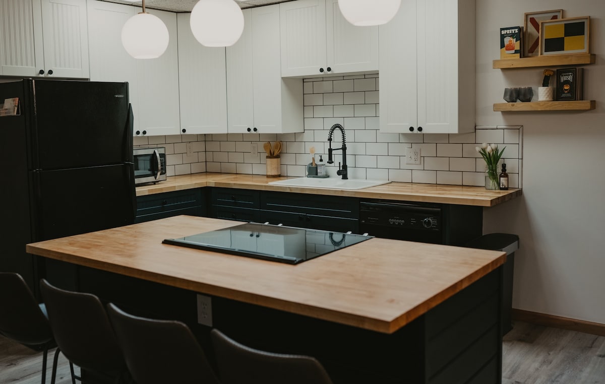 The kitchen features a centered wooden island equipped with a stovetop. Cabinets are arranged above and below a tiled backsplash. A black refrigerator and microwave are visible, along with minimal decor on open shelving. Warm overhead lighting brightens the space.