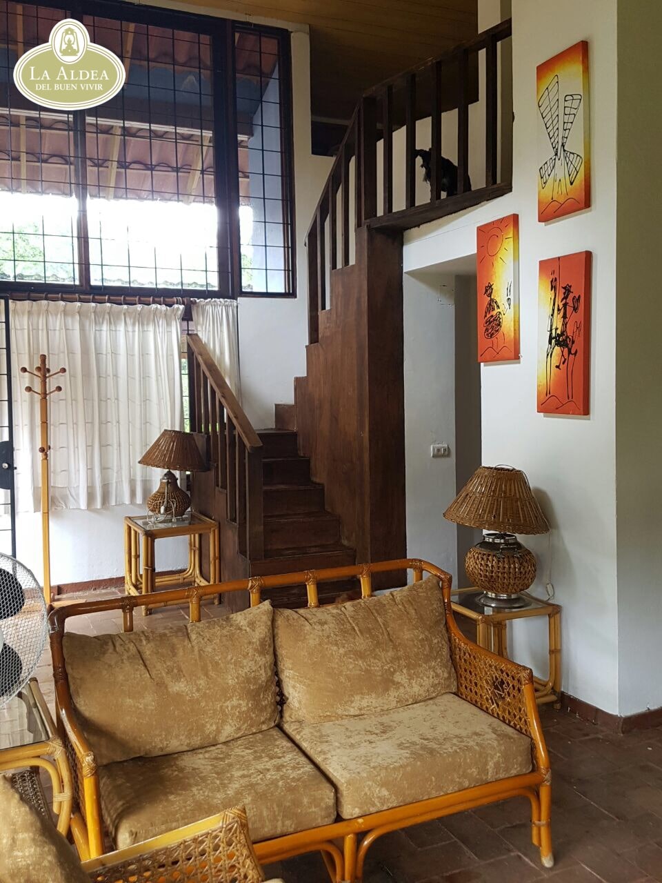 A cozy living area is furnished with soft golden-brown seating and complemented by two decorative lamps. A wooden staircase leads to the upper level, framed by large windows that allow natural light to fill the space. Artwork is displayed on the walls, adding color.