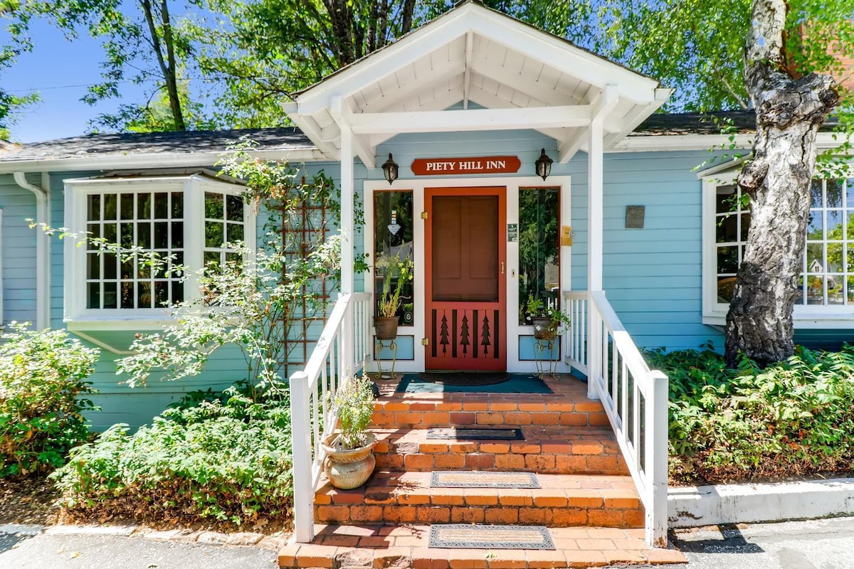 The exterior of Piety Hill Inn is showcased, featuring a welcoming entrance with a red door framed by white trim. Lush greenery and potted plants line the steps, while windows allow natural light to illuminate the space. A charming pathway leads toward the inviting entryway.