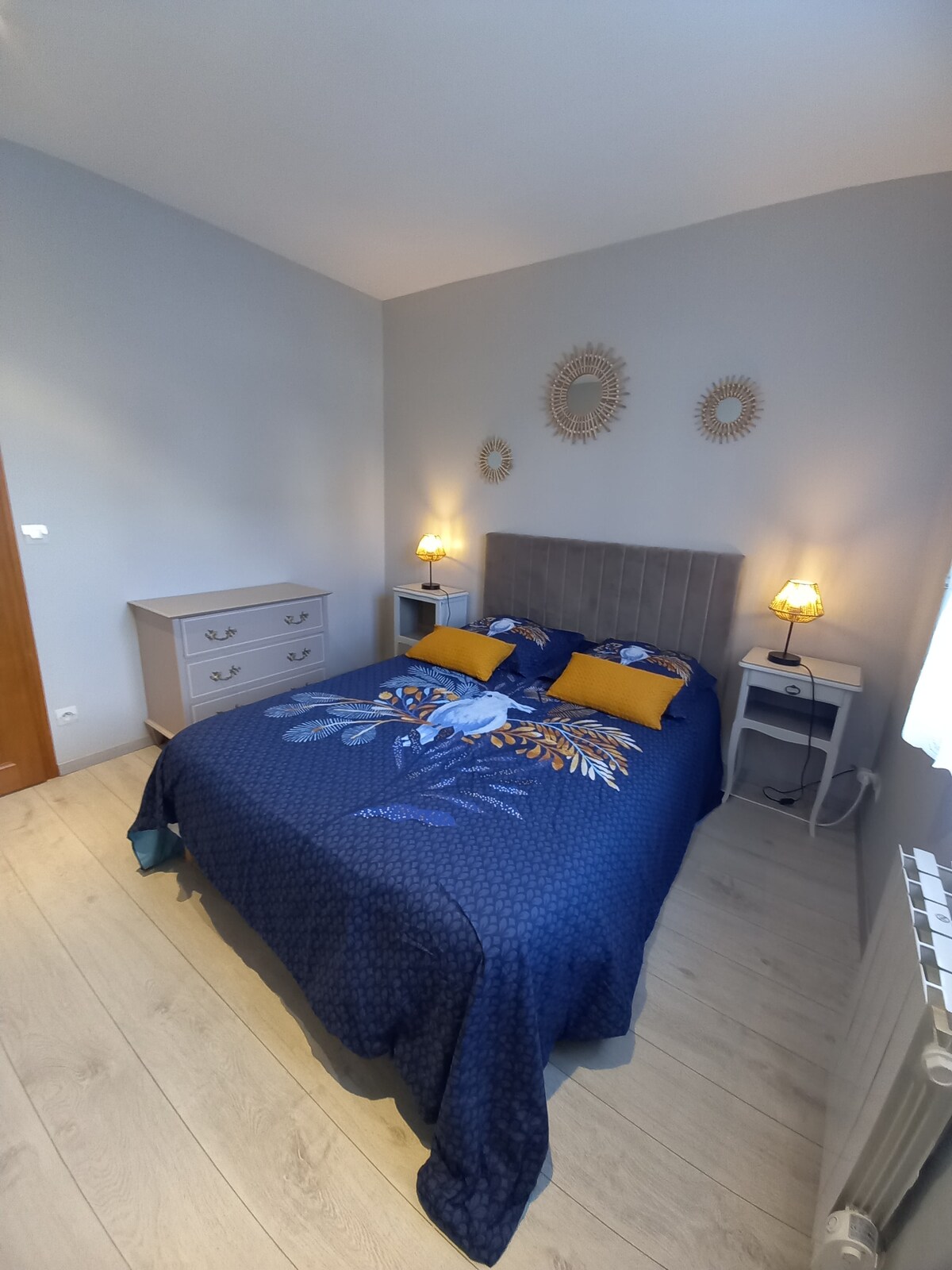 A bright bedroom features a spacious bed adorned with blue and yellow accent pillows and a patterned comforter. Soft lighting from a bedside lamp creates a cozy atmosphere. A dresser stands in the corner, complemented by a simple nightstand beside the bed.
