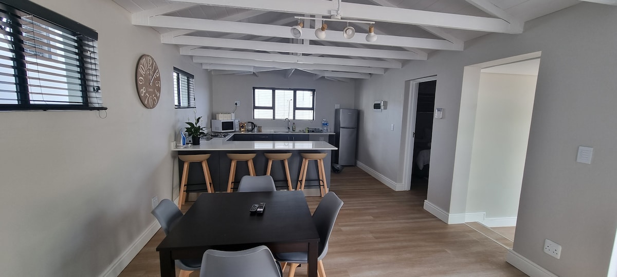 A spacious open-plan kitchen and dining area feature a large island with high stools and a wooden dining table surrounded by chairs. Natural light filters through the window treatments, highlighting the sleek design of the space. Modern appliances are visible in the kitchen area.