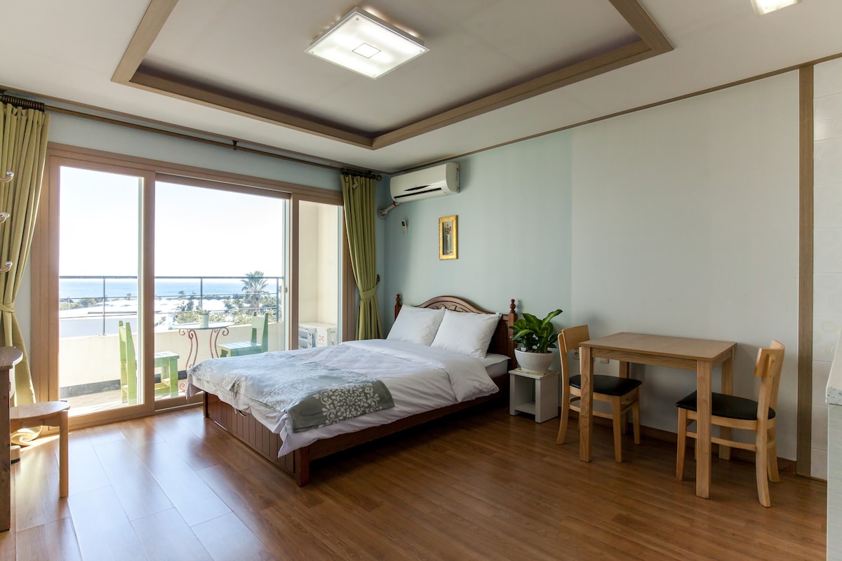A bright bedroom features a spacious bed with crisp white linens. Large sliding glass doors offer a view of the sea and provide natural light. A small wooden table with two chairs and a potted plant are positioned near the window, enhancing the serene atmosphere.