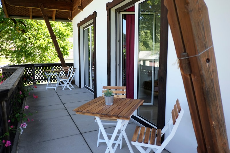 An outdoor balcony features a wooden table surrounded by two white chairs. Large glass doors provide access to the interior, with leafy greenery partially visible beyond. The flooring is tiled, and decorative flowers add a touch of color to the space.