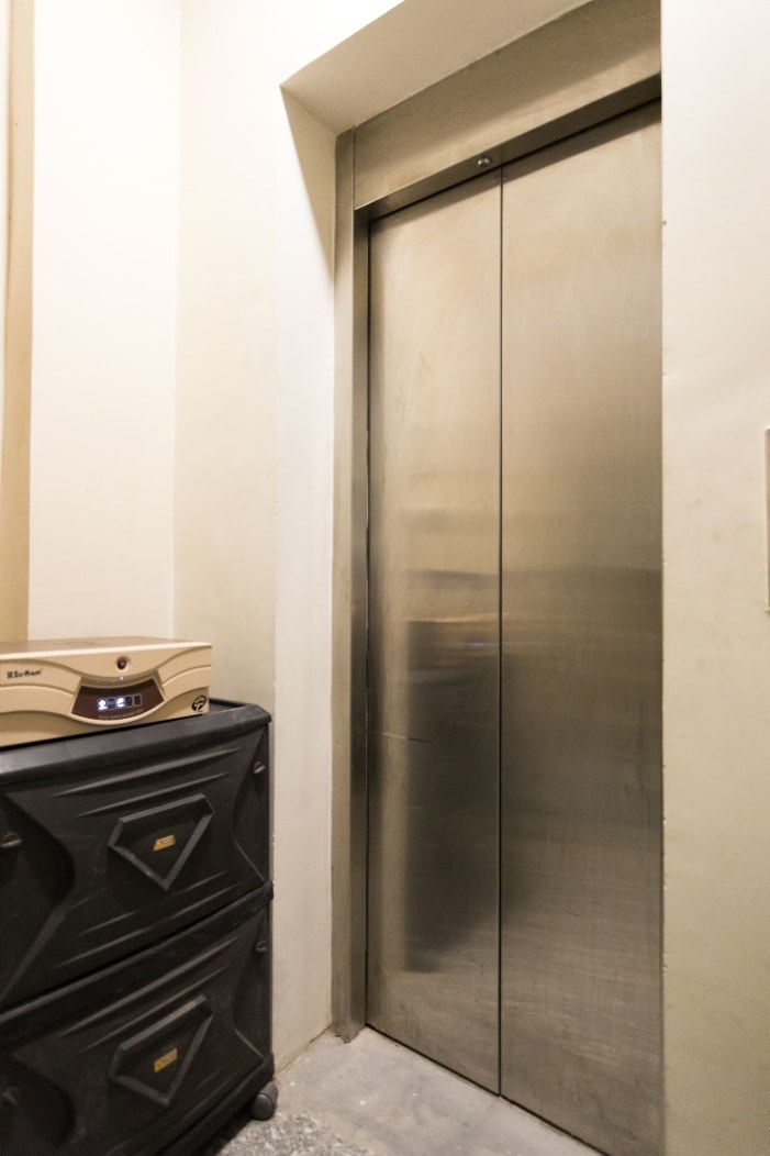 A metal elevator door stands closed within a narrow hallway. Adjacent to the elevator, a small cabinet can be seen, providing additional storage. The surroundings feature a polished concrete floor and light-colored walls, highlighting a simple and functional design.