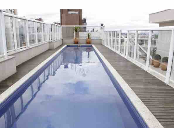 A rooftop pool is surrounded by glass barriers, offering a clear view of the city skyline. The pool's still water reflects the sky, while potted plants add touches of greenery along the deck.