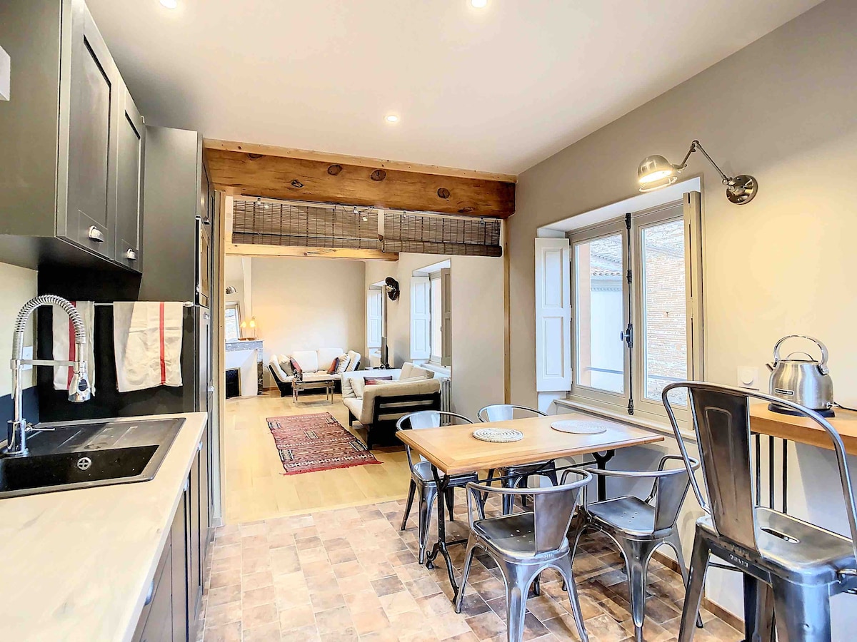 A well-equipped kitchen area features dark cabinetry and a dining space with a wooden table. Industrial-style chairs complement the setting. Natural light enters through multiple windows, creating a warm ambiance. The living area is visible in the background, showcasing a comfortable seating arrangement.