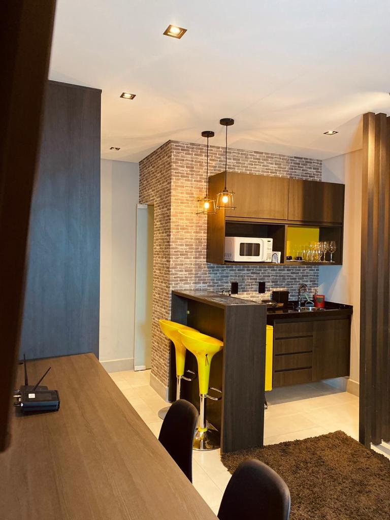 A modern kitchen area is presented, featuring a sleek design with dark cabinetry and yellow accent stools. A textured brick wall offers visual interest. Essential appliances, including a microwave and coffee maker, are easily accessible, complemented by organized kitchenware and a well-lit atmosphere.