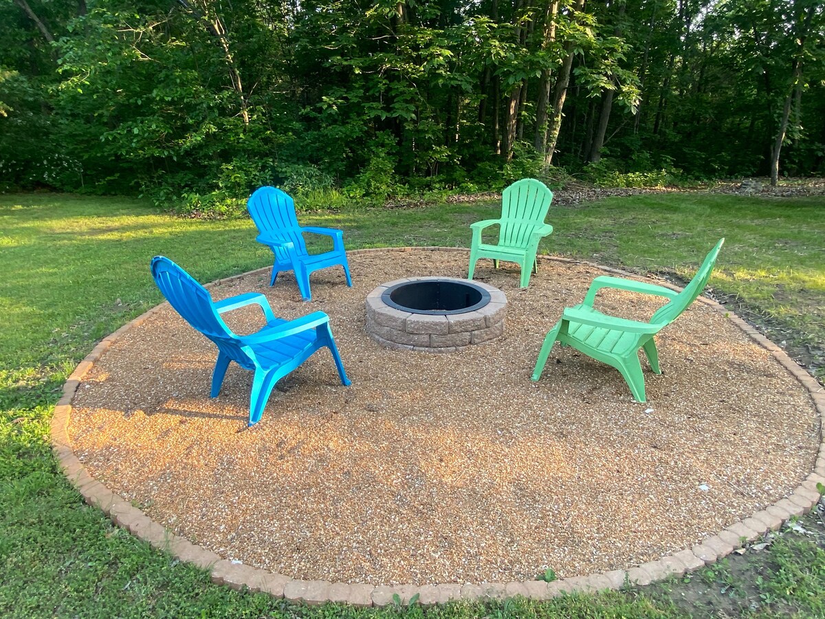 A circular firepit area is surrounded by gravel, featuring four colorful Adirondack chairs in blue and green. Lush greenery from trees provides a natural backdrop, creating a serene setting for outdoor gatherings.
