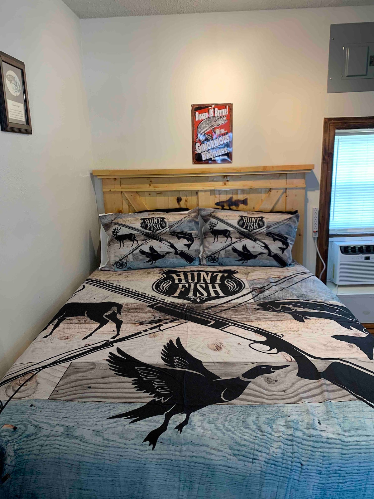 A sleeping area features a wooden headboard and a bed adorned with a hunting and fishing themed quilt, displaying black illustrations of birds and fishing gear. Two matching pillows complete the bedding. A window with light filtering through is situated nearby.