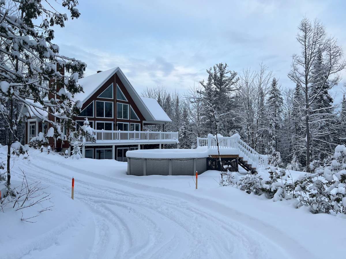A chalet is surrounded by a winter landscape, with snow blanketing the ground and trees. The property features a large deck with white railings and a slope leading to the entrance. An outdoor hot tub is positioned along the deck's edge, offering a serene view.