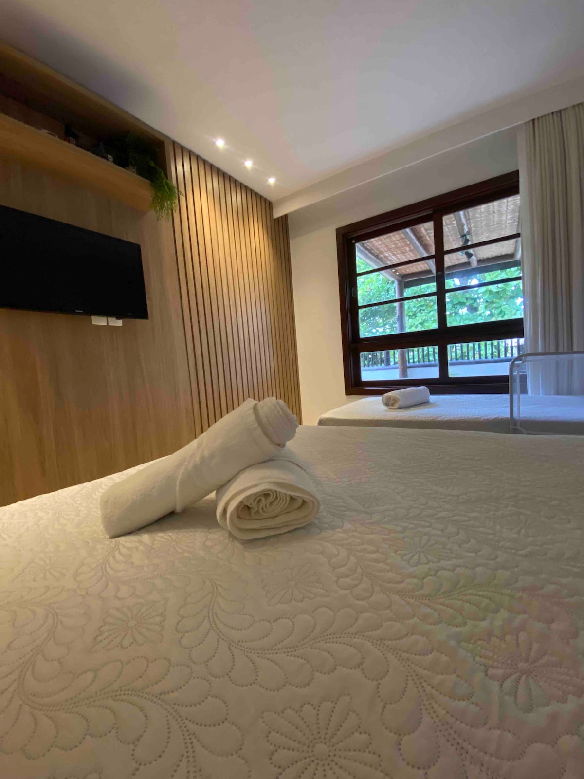 A light and airy bedroom is depicted, featuring a spacious bed with a textured quilt. Rolled towels are neatly placed on top. A large window invites natural light and overlooks a balcony with wooden details. A flat-screen television is mounted on the wooden wall.