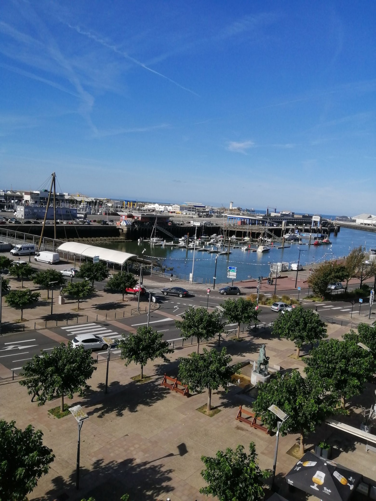 A scenic view of the harbor is captured, featuring calm waters and moored boats. Lush trees line the waterfront promenade, while vehicles pass along the street. The clear blue sky enhances the tranquil atmosphere of the port area.
