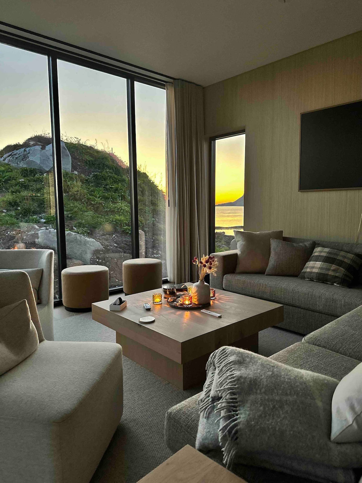 A modern living area features large glass windows showcasing a sunset view over the landscape. Comfortable seating includes soft couches and stools arranged around a wooden coffee table. Minimal decorative elements create a serene atmosphere, complemented by natural light filtering through sheer curtains.
