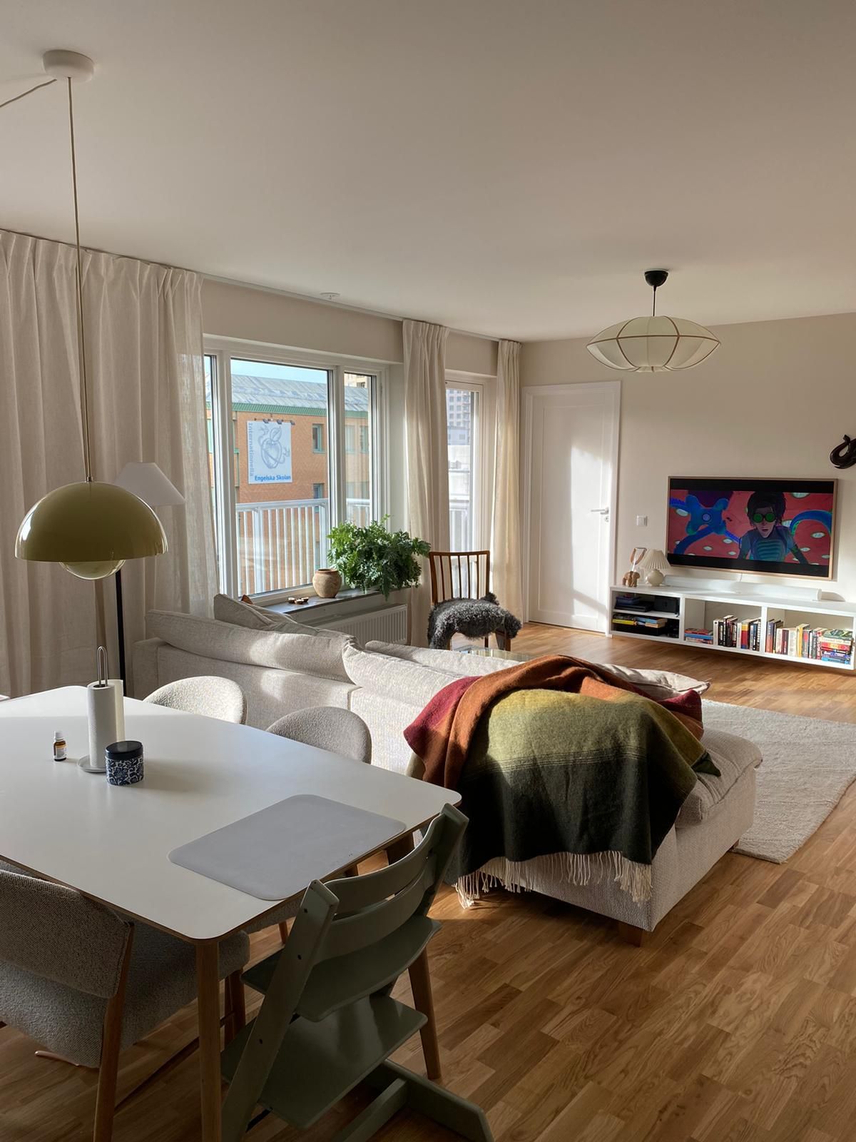 A bright living area is shown, featuring large windows with sheer curtains allowing natural light to enter. A comfortable sofa is present, adorned with a colorful blanket draped over the arm. A dining table set for four is positioned alongside a stylish lamp, viewing a television mounted on the wall.