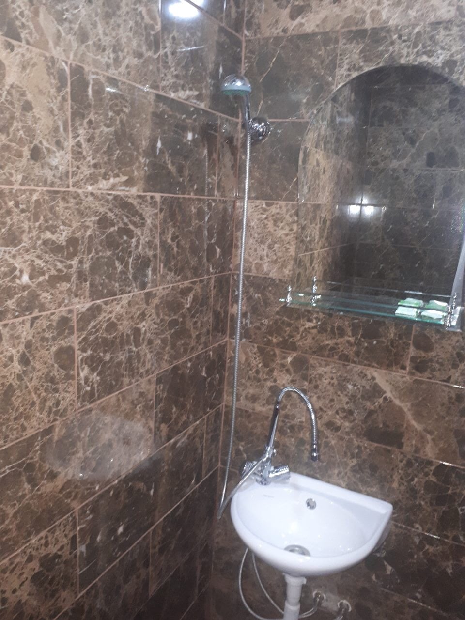 A modern bathroom features stylish marble walls, creating a rich texture. A compact sink is positioned below a wall-mounted mirror, while a showerhead is mounted above. The overall design offers a clean and functional space for privacy and comfort.