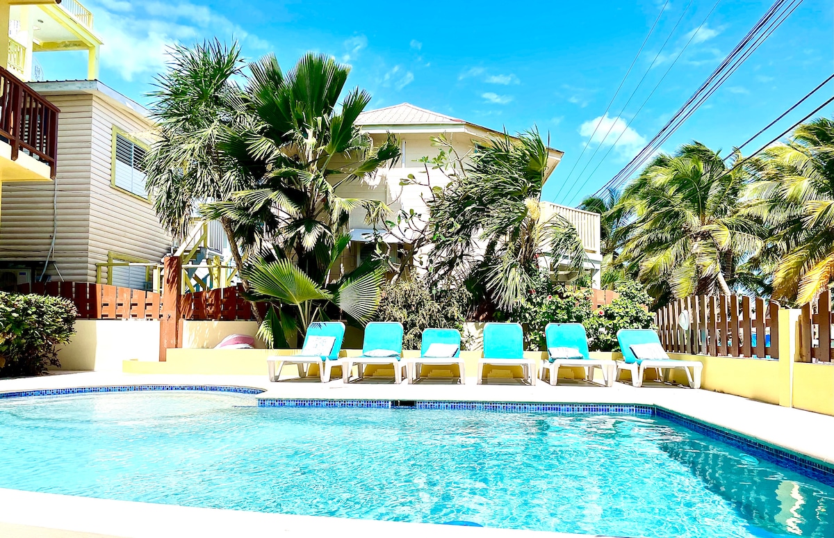 A refreshing swimming pool is bordered by lush tropical plants and features multiple lounge chairs arranged around its inviting perimeter. The building in the background adds to the serene setting, with blue skies overhead and gentle movement from palm trees in the breeze.