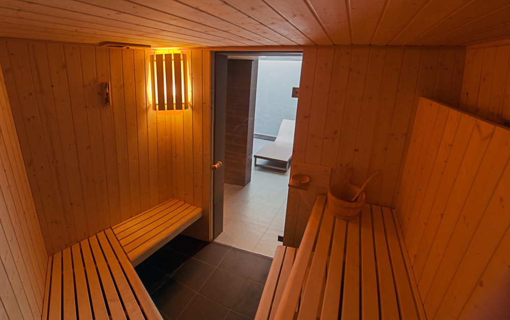 A wooden sauna features horizontal bench seating along the walls, with soft lighting creating a warm glow. A small bucket is visible for water use, and a door leads to a relaxing area with lounge chairs. The flooring is finished in dark tiles.