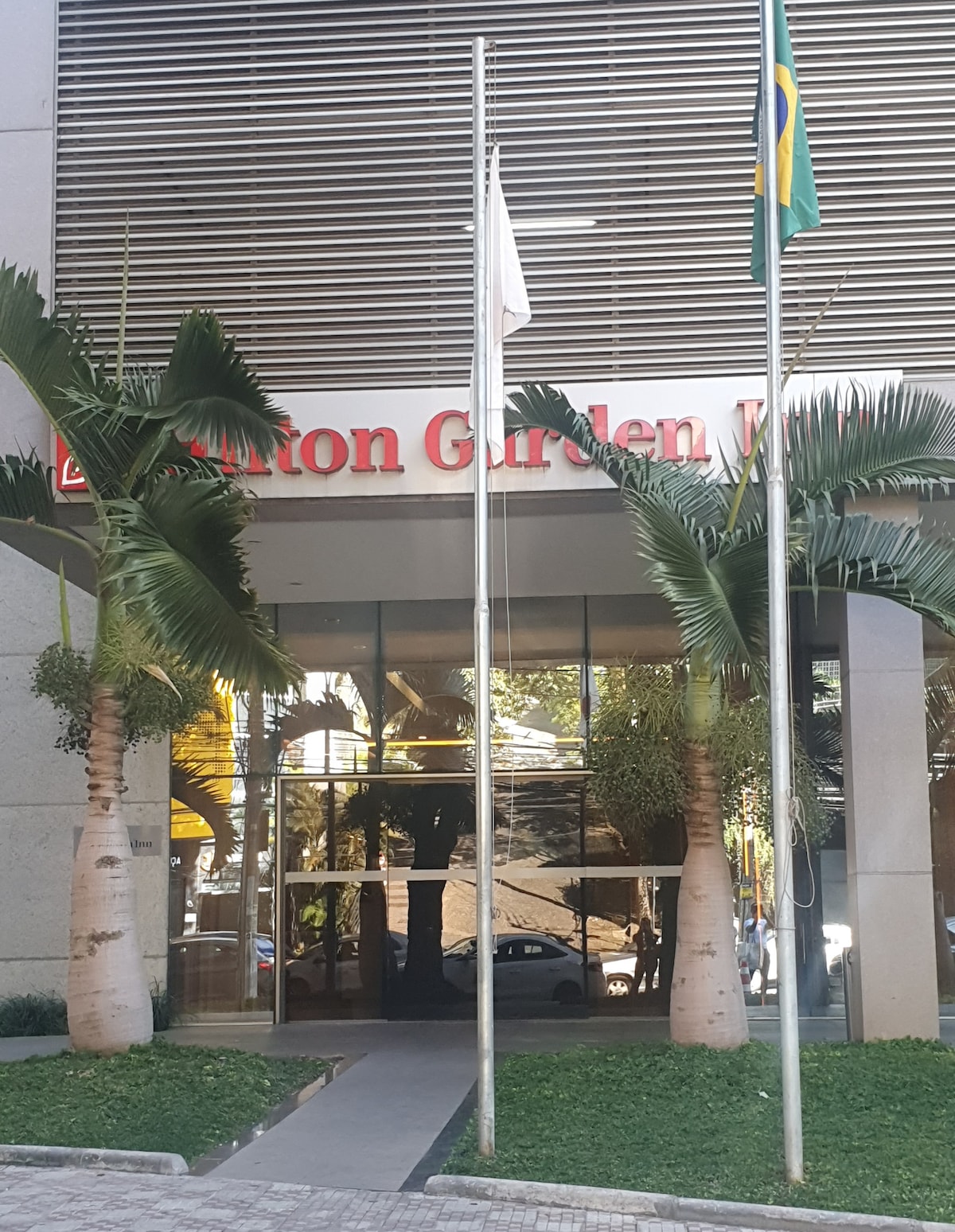 The entrance of the Hilton Garden Hotel is framed by tall palm trees. A welcoming atmosphere is created by the modern glass doors, which reflect the surrounding area. The hotel's name is prominently displayed above the entrance in bold letters.