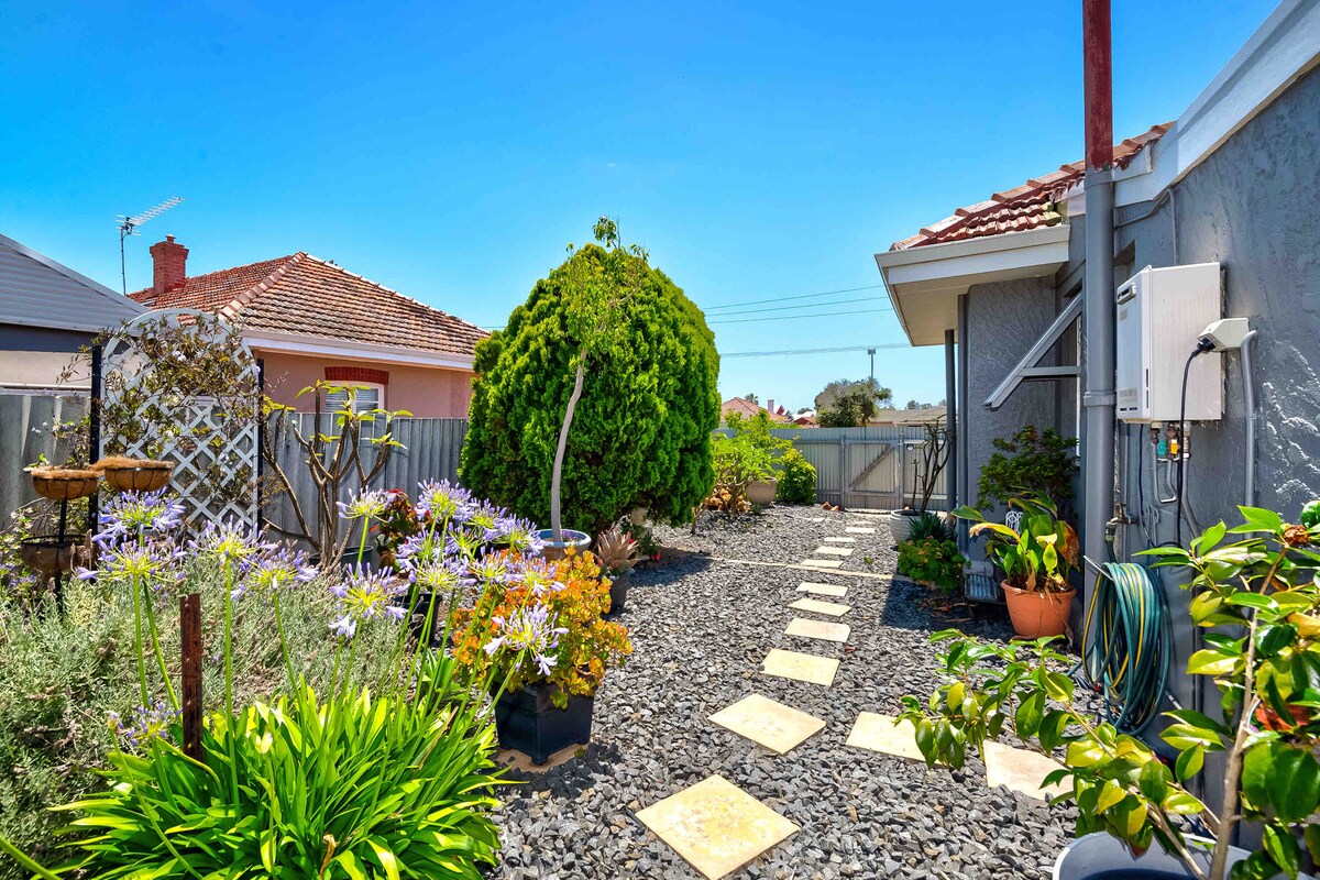 A serene garden pathway is bordered by a variety of plants and flowers, including vibrant green shrubs and flowering bushes. Stone stepping stones lead through the garden, which features decorative planters and a gravel surface. The area is enclosed with a fence, providing privacy.